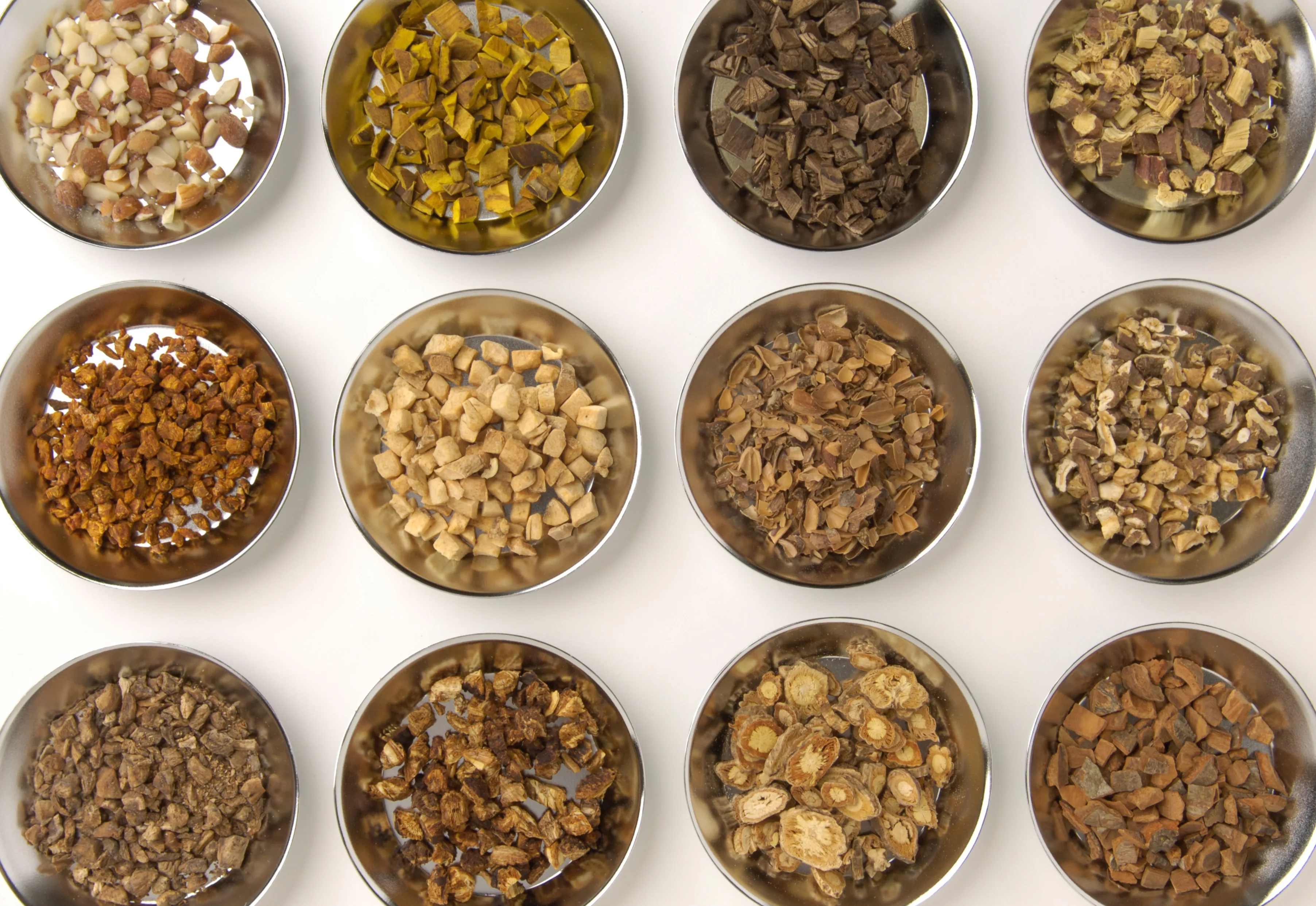 Twelve polished dishes of dried Chinese herbal granules arranged on white